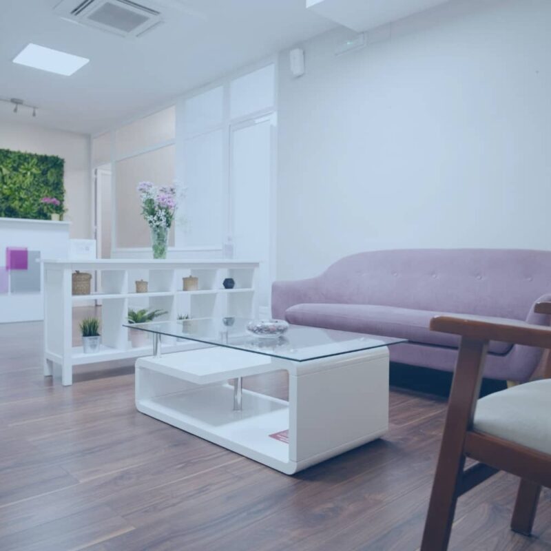 A modern, minimalist wellness room featuring a lavender sofa, dark wood floors, and a vertical green plant wall behind a white reception desk.