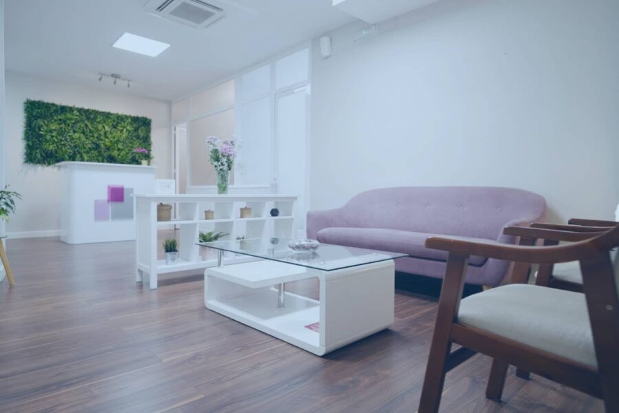 A modern, minimalist wellness room featuring a lavender sofa, dark wood floors, and a vertical green plant wall behind a white reception desk.