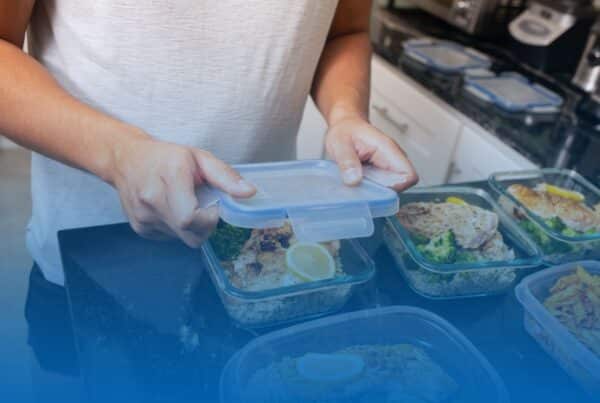 A person placing a lid onto a glass meal prep container filled with grilled chicken, broccoli, and rice on a kitchen counter.