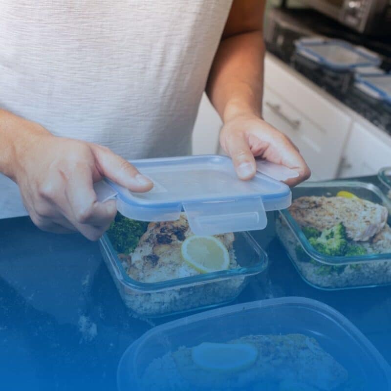 A person placing a lid onto a glass meal prep container filled with grilled chicken, broccoli, and rice on a kitchen counter.