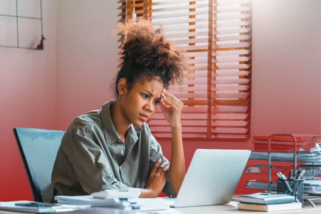 A stressed young woman sitting at her office desk, resting her hand against her head while looking intently at a laptop screen.