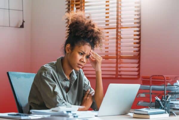 A stressed young woman sitting at her office desk, resting her hand against her head while looking intently at a laptop screen.