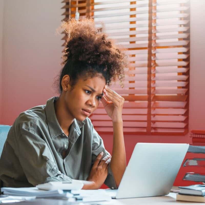 A stressed young woman sitting at her office desk, resting her hand against her head while looking intently at a laptop screen.