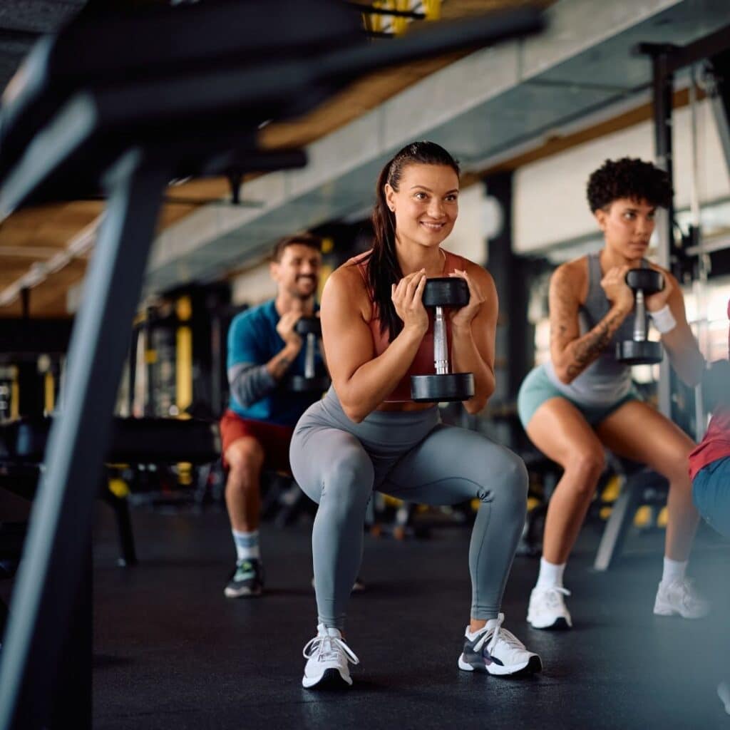 A smiling woman holding a dumbbell while performing a goblet squat during a group fitness class at a gym.