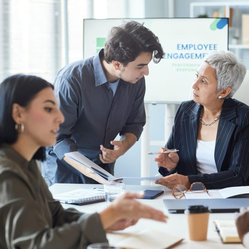 A human resources manager reviewing documents with colleagues in a office meeting with an employee engagement presentation in the background.