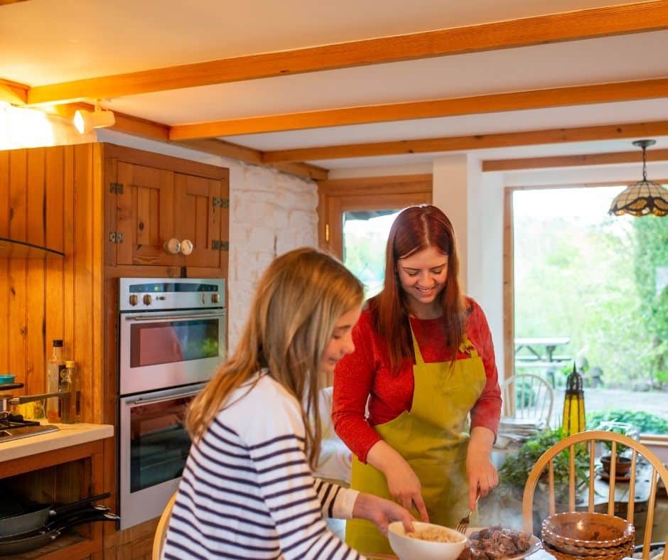 Two women preparing a meal together in a cozy kitchen, with one in a green apron slicing food while the other handles a bowl at the table.