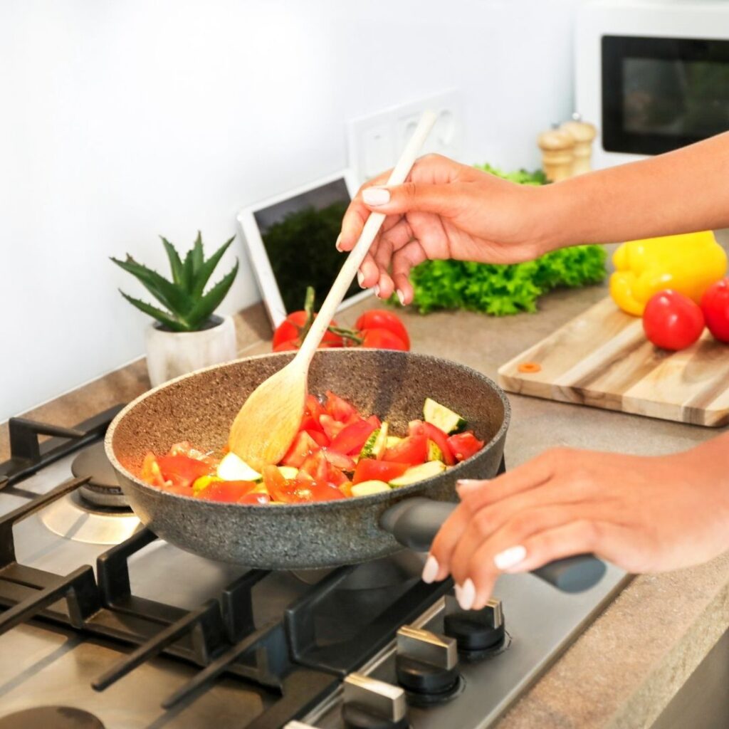 A person stirring sliced tomatoes and zucchini in a skillet on a gas stove with a wooden spoon, with fresh peppers and produce on the kitchen counter.