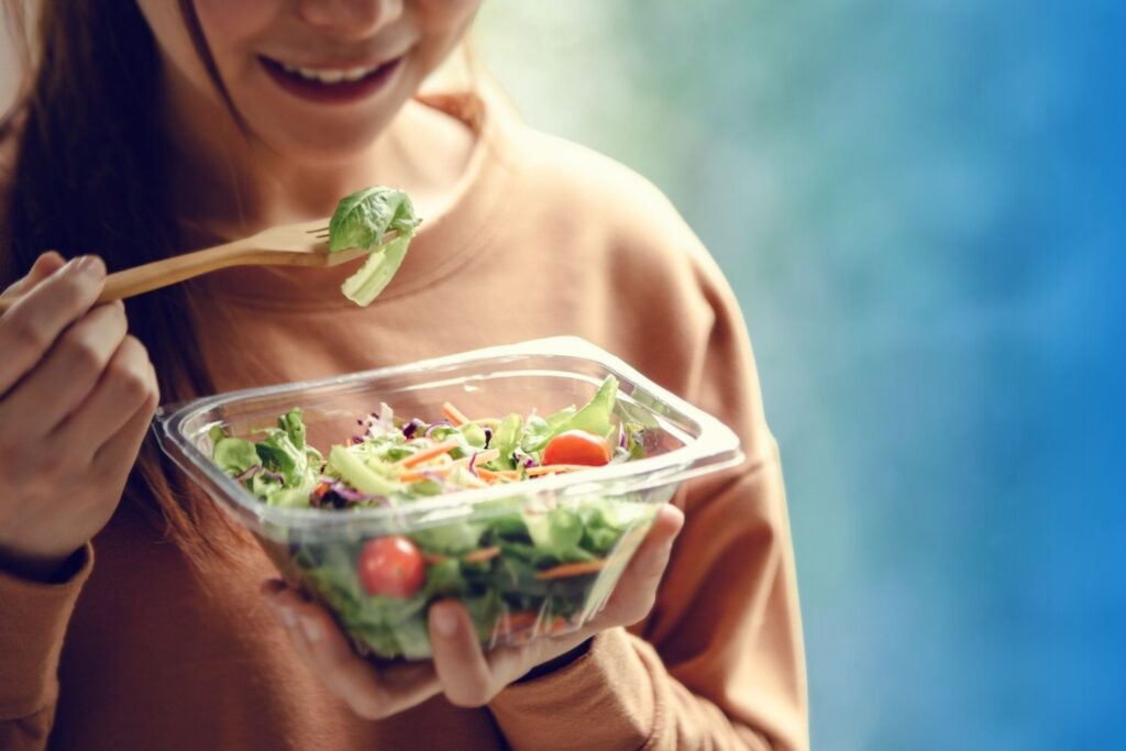 A person holding a clear plastic container of fresh green salad with cherry tomatoes and eating with a wooden fork.