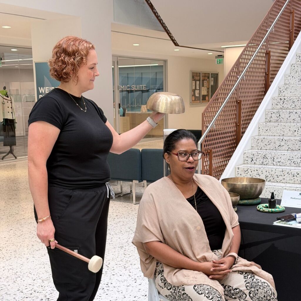 A wellness practitioner holds a brass singing bowl above a seated attendee's head during a sound therapy demonstration at an indoor health fair.